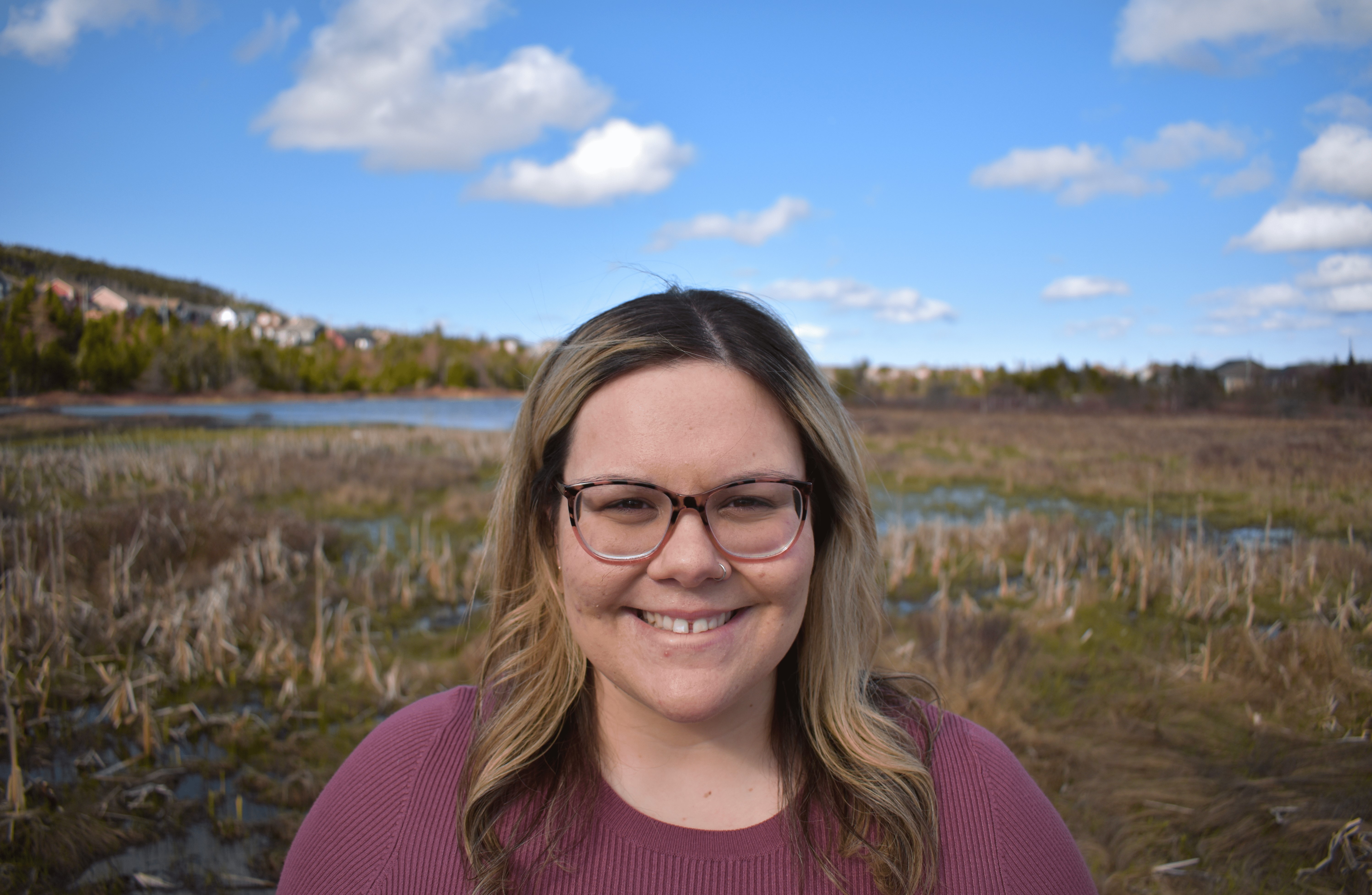 A young blonde woman wearing glasses stands in front of a field with a pond in the background and blue skies